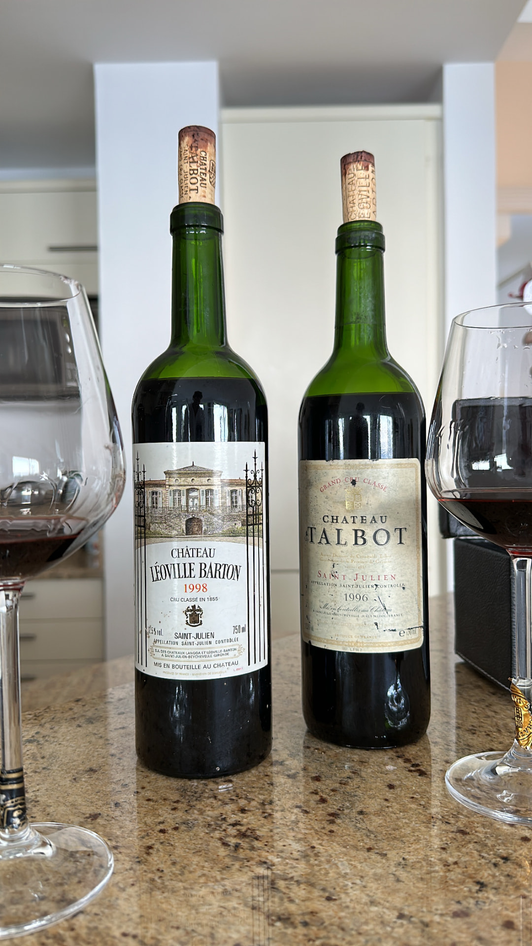 Bottles of 1998 Château Léoville Barton and 1996 Château Talbot standing between two glasses of red wine on a kitchen counter.